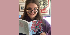 Jessica Watkin holds a copy of her book, Interdependent Magic. She has long brown hair, and wears glasses and a purple sweater.