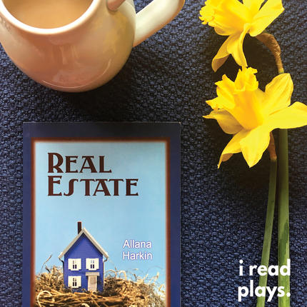 A copy of Real Estate lies on a table beneath a mug and next to flowers
