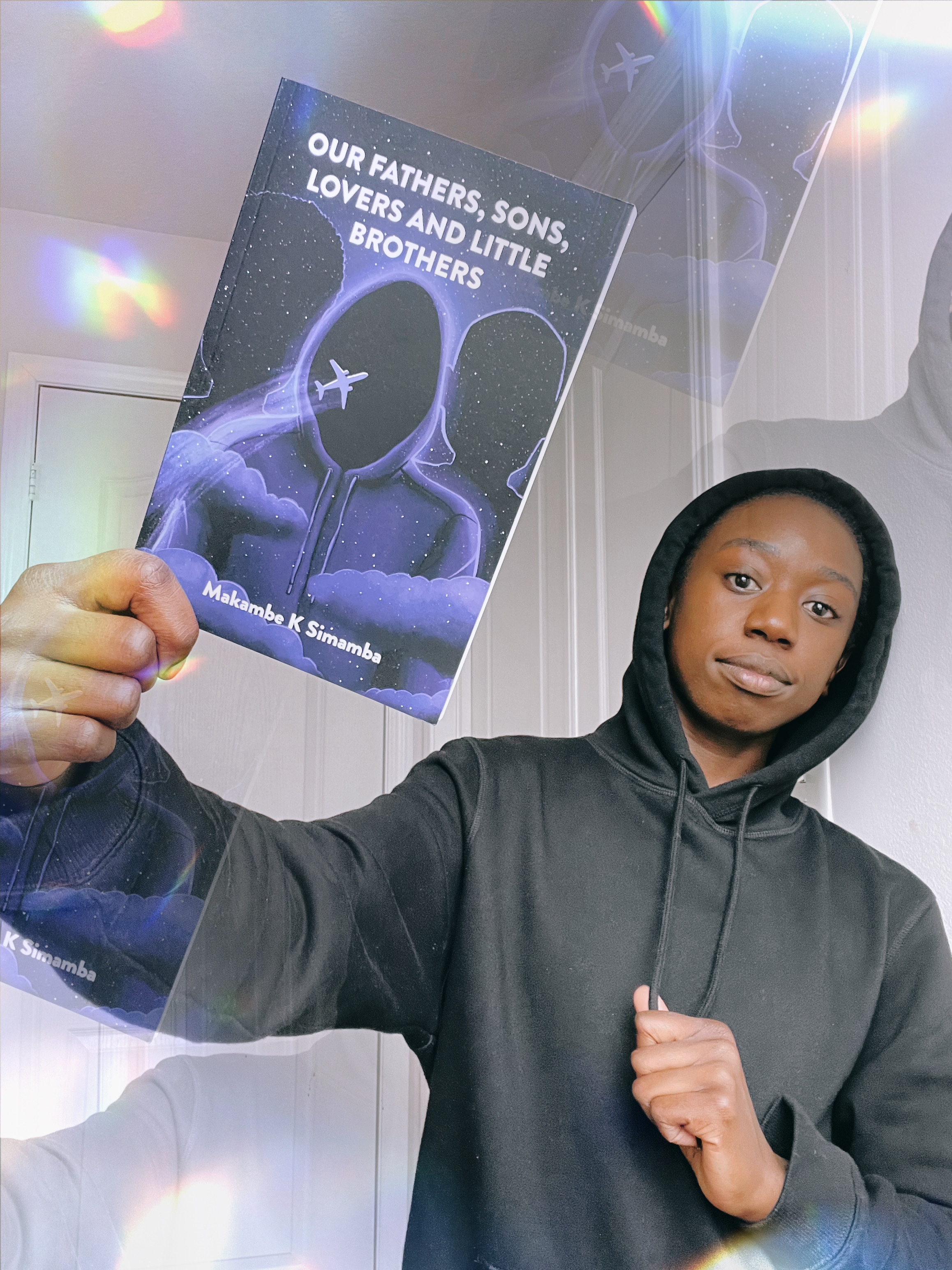 Makambe K Simamba dressed in character, wearing a black hoodie, for Our Fathers, Sons, Lovers and Little Brothers, holds a copy of the book